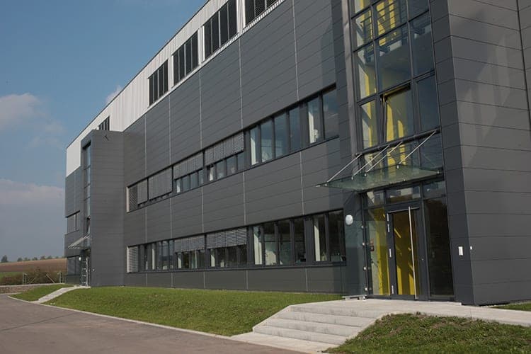 Eingang zum Bürotrakt mit gläsernem Vordach, Treppenstufen und gelber Wand hinter der Glasfassade
