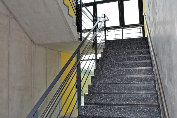 Treppenhaus mit Sichtbetonwänden, gesprenkelten Stufen, Stahlgeländer und gelber Wandfläche