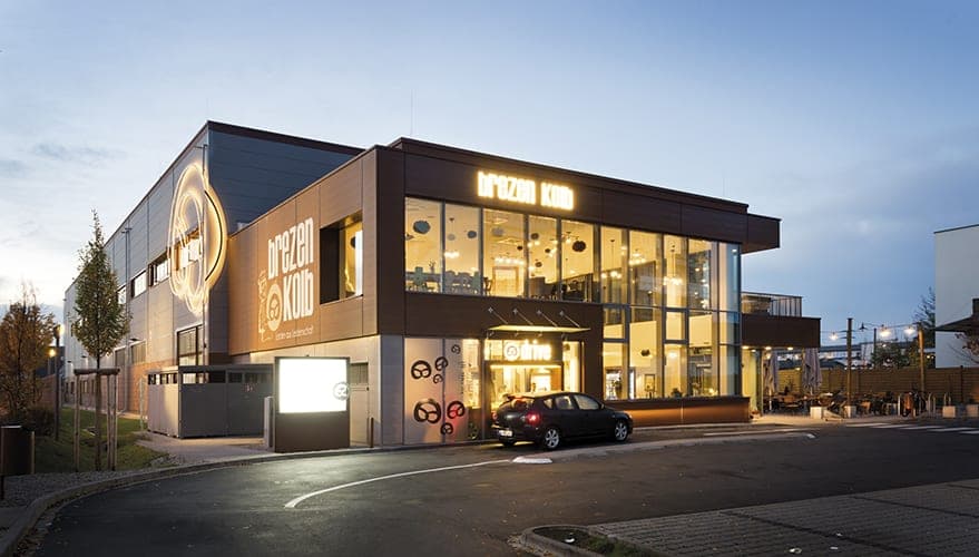 Glazed entrance front with illuminated lettering and a drive-through counter at dusk