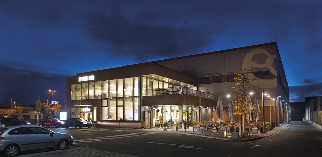 Building corner at night with the lit café, outdoor terrace and round logo on the hall façade