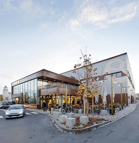 Glazed building corner with a roof terrace above the café and a forecourt with concrete stools by day