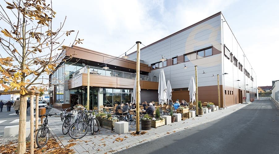 Café terrace with guests, planters and bicycle parking in front of the hall façade