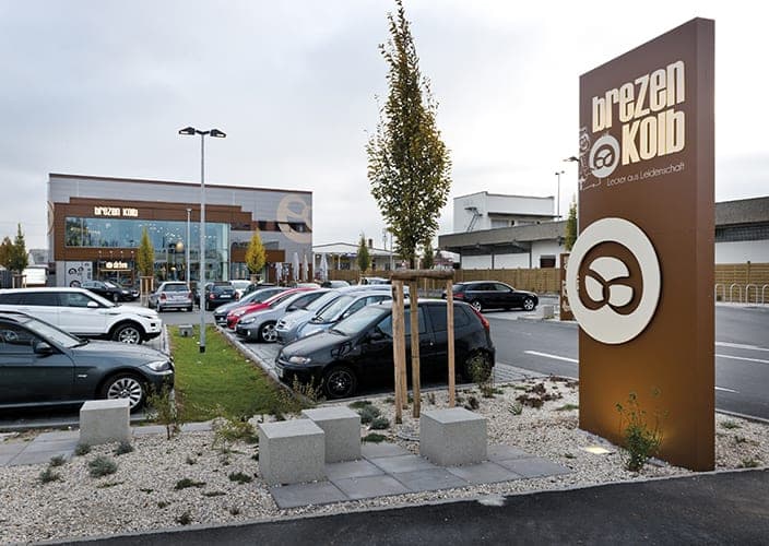Car park with a free-standing logo pylon, concrete stools and trees, the café beyond