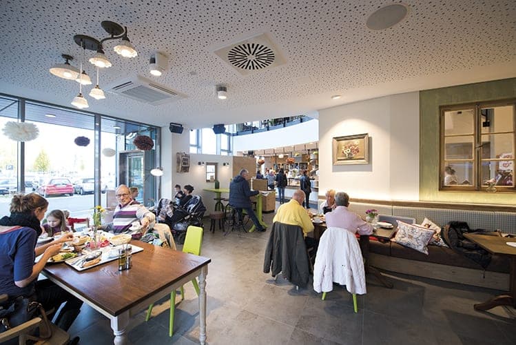 Café seating with guests at wooden tables, a bench seat and a perforated acoustic ceiling