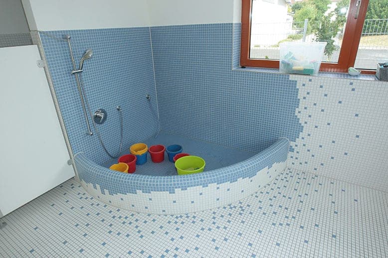 Blue-tiled splash area with a hand shower, mosaic gradient on the floor and colourful play buckets