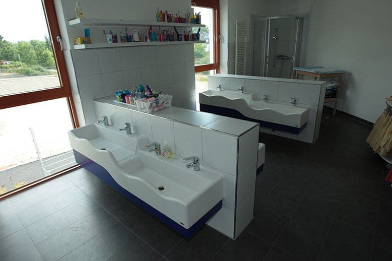 Washroom with two curved rows of washbasins, toothbrush cups on wall shelves and a shower behind