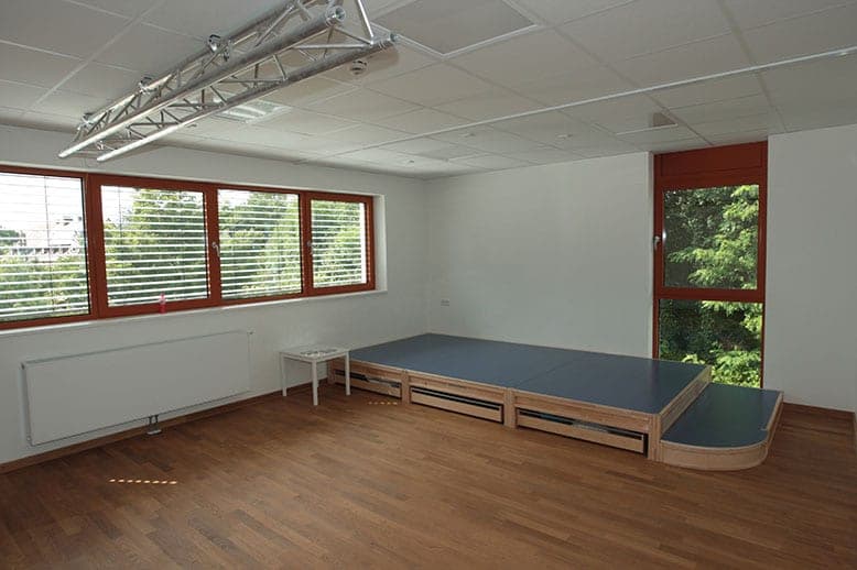 Room with a timber stage platform, parquet floor, lighting truss and windows looking out onto trees