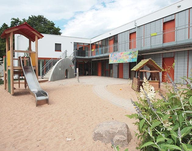 Courtyard with a sand play area in front of the two-storey building with access gallery and orange doors