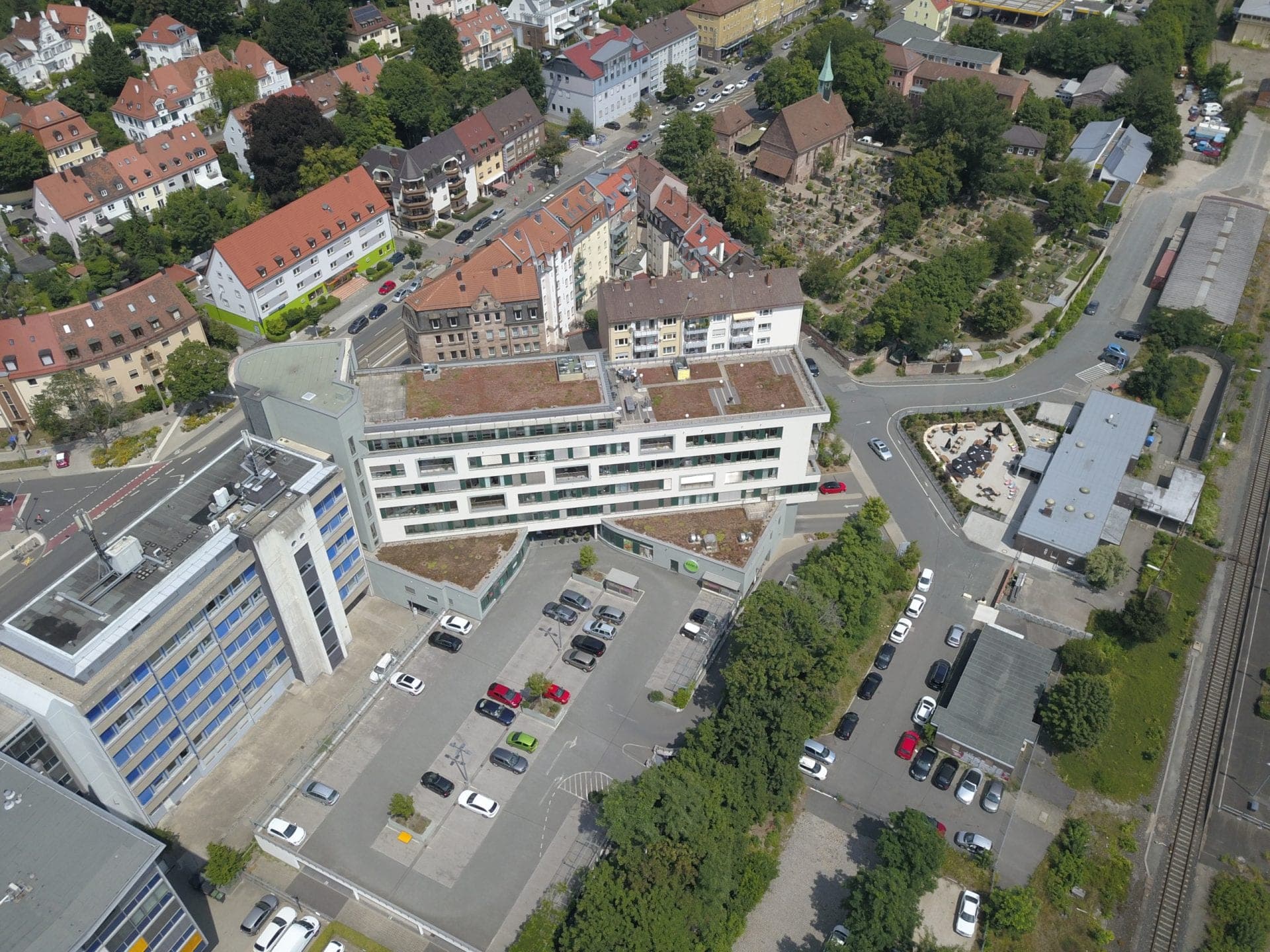 Luftbild des Gebäudekomplexes mit begrünten Dächern und Parkplatz im Hof, im Hintergrund Kirche und Friedhof