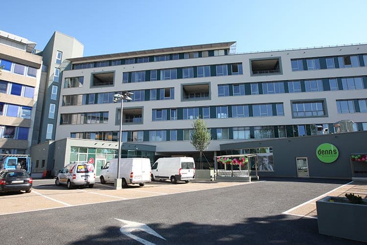 Frontale Ansicht der Hoffassade mit Loggien, davor Parkplatz, Lichtmast und eingeschossiger Ladenbau