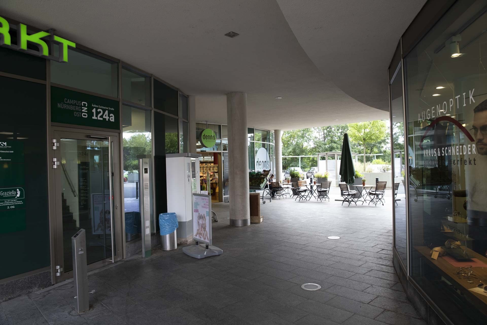 Überdachter Durchgang mit Ladenfronten, Hauseingang und Außensitzplätzen vor dem Biomarkt