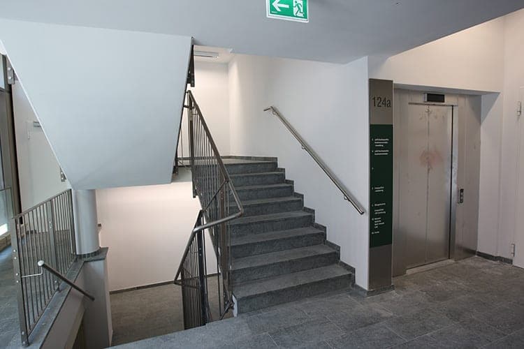 Treppenhaus mit Natursteinstufen, Edelstahlhandlauf, Aufzug und Wegweiser im Erschließungsbereich