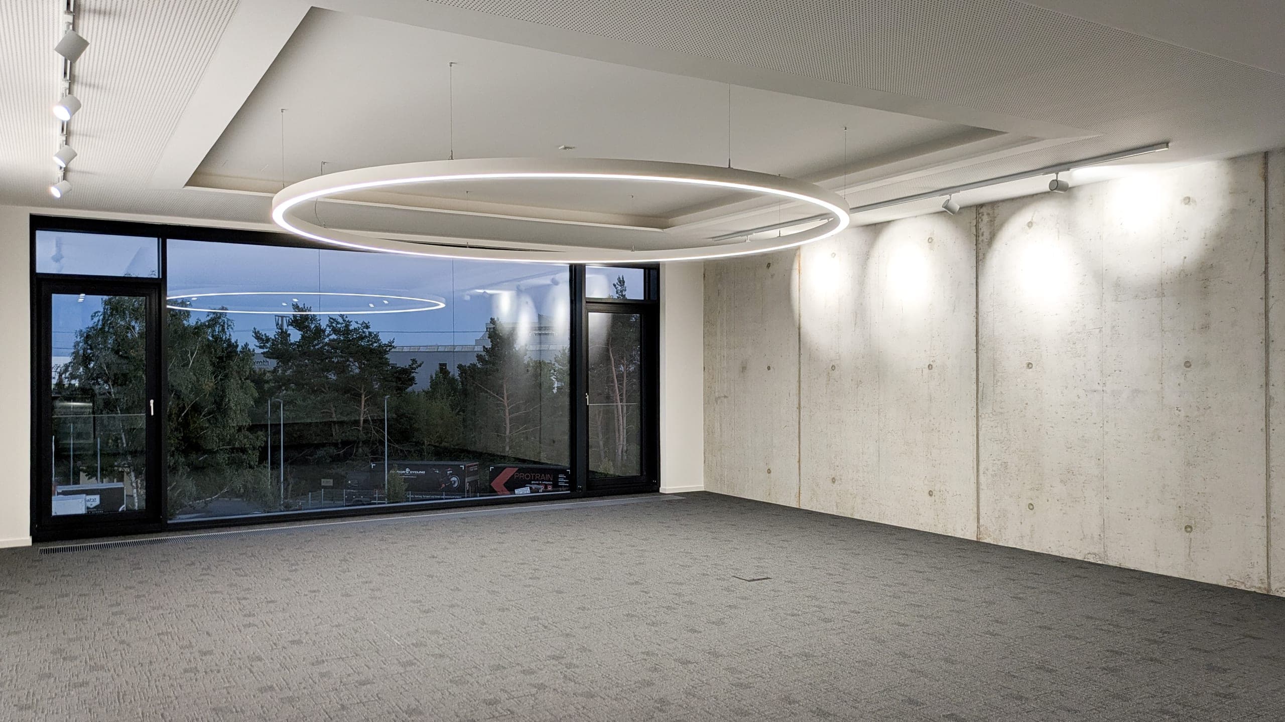 Leerer Raum mit ringförmiger Pendelleuchte, Sichtbetonwand und raumhoher Fensterfront in der Dämmerung