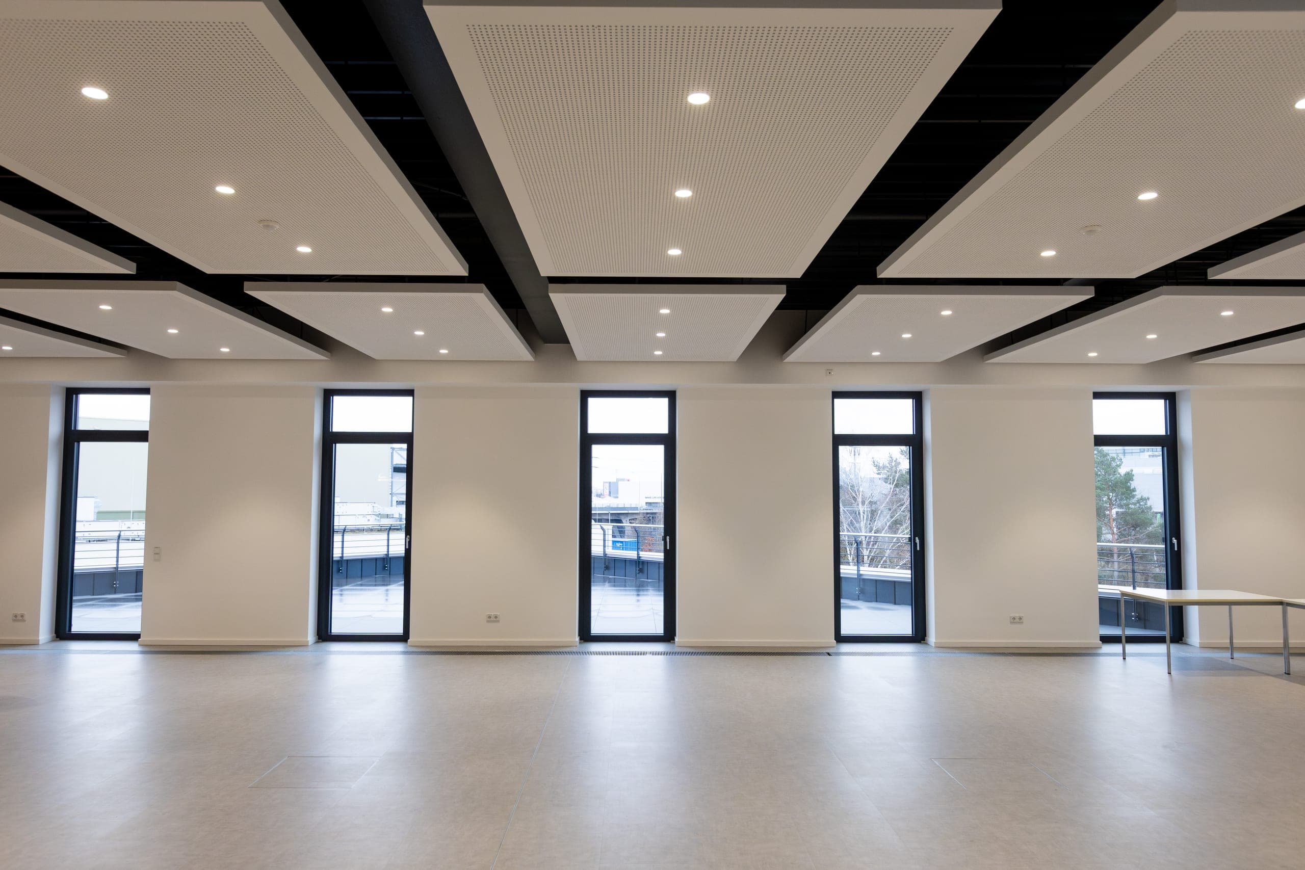 Heller Saal mit fünf raumhohen Fenstertüren zur Dachterrasse und Akustiksegeln an der Decke
