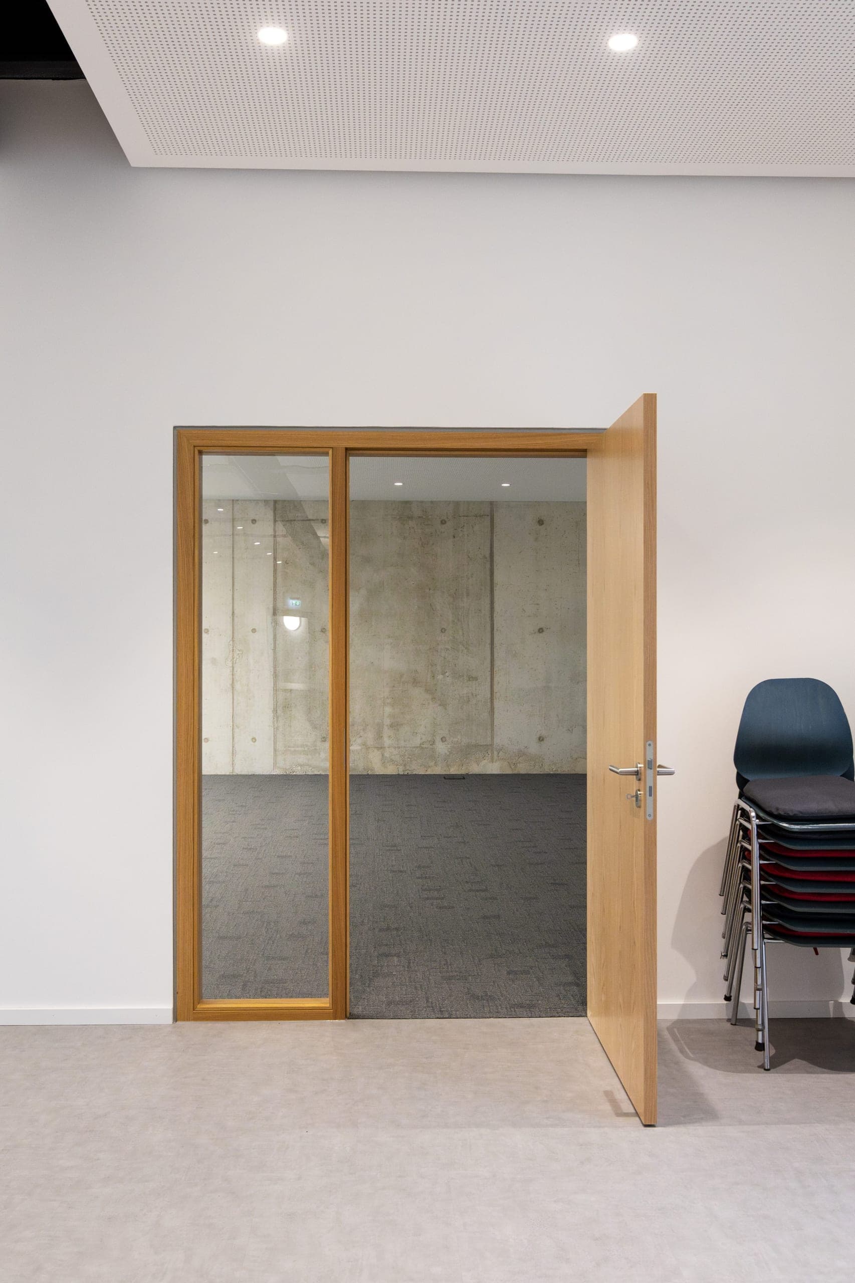 Blick durch eine offene Holztür mit Glasseitenteil in einen leeren Raum mit Sichtbetonwand