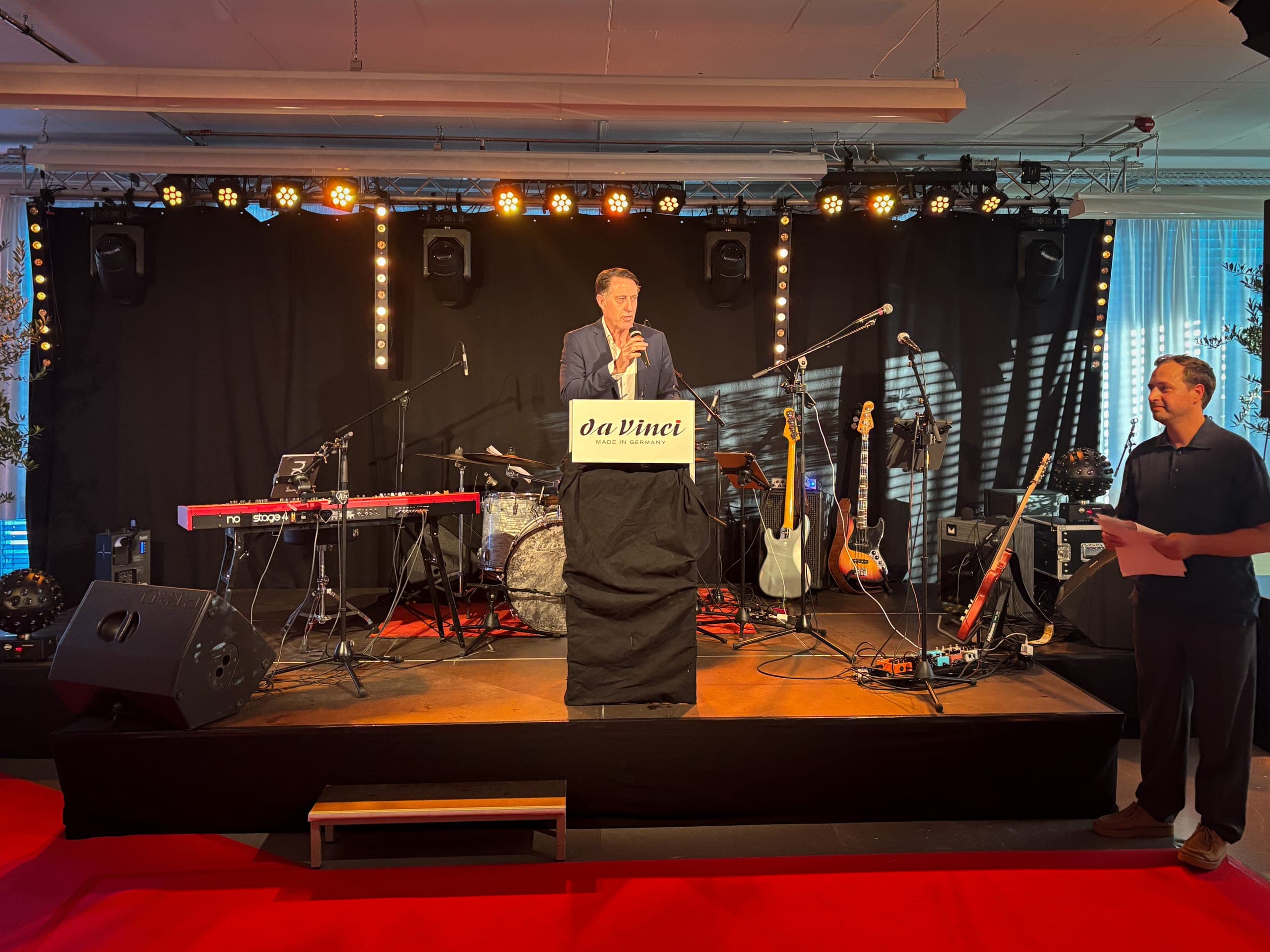Rede am Rednerpult mit daVinci-Schild bei einer Firmenveranstaltung, dahinter eine Bühne mit Bandinstrumenten