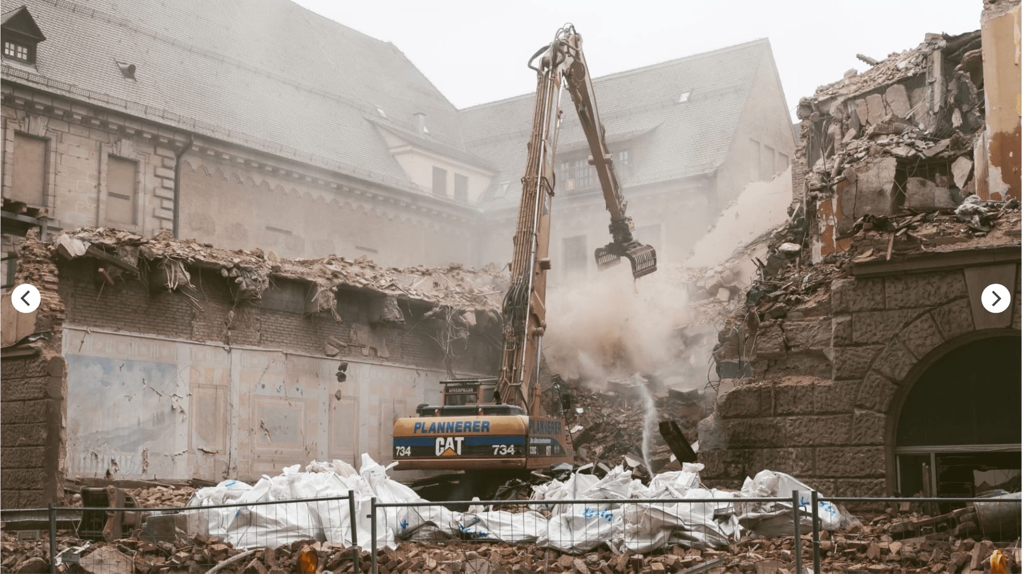 Demolition excavator taking down parts of the building, with dust over rubble and filled bags in front of the old building