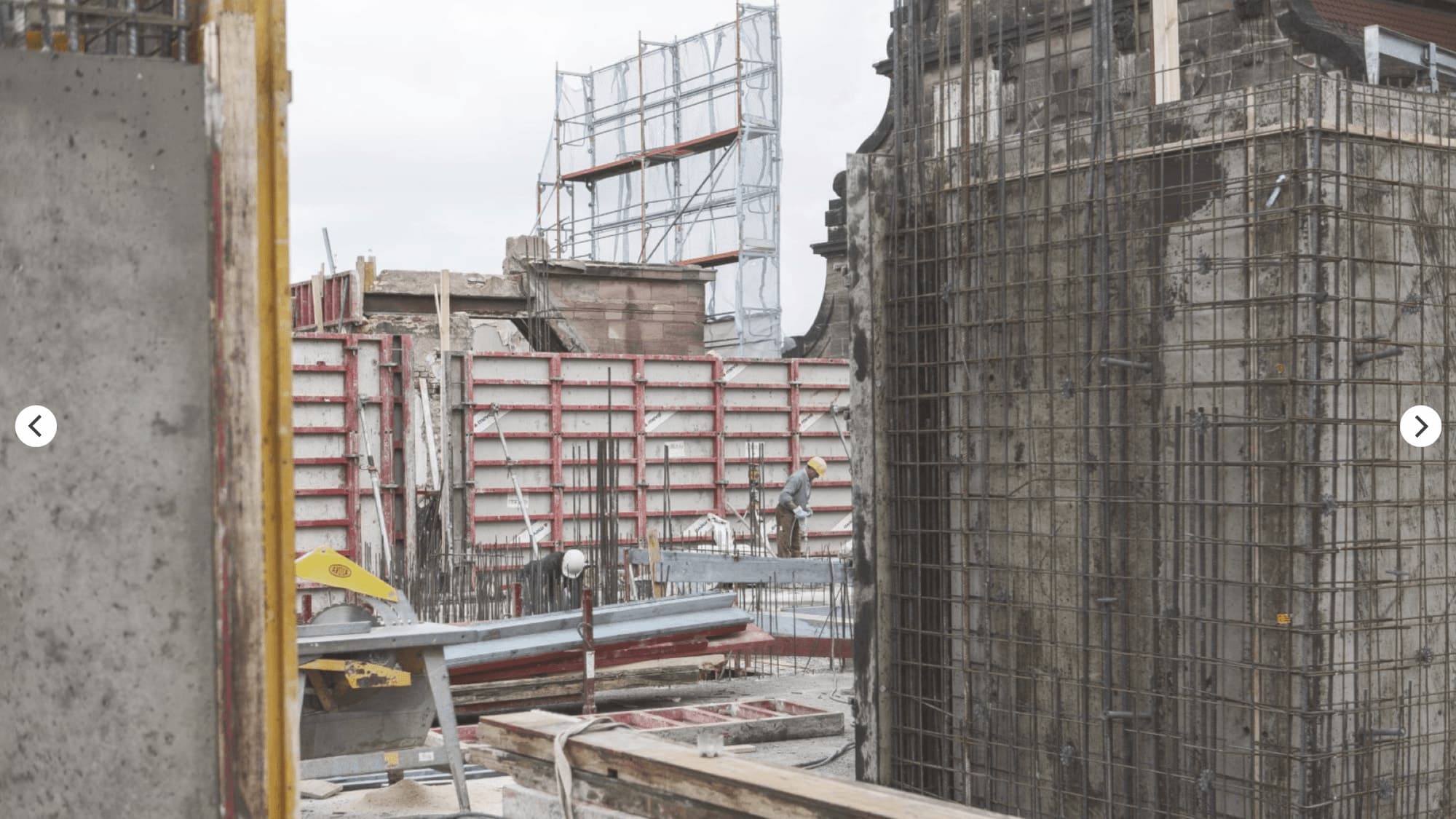 Construction site with formwork panels, workers, reinforcement on an existing wall and scaffolding behind