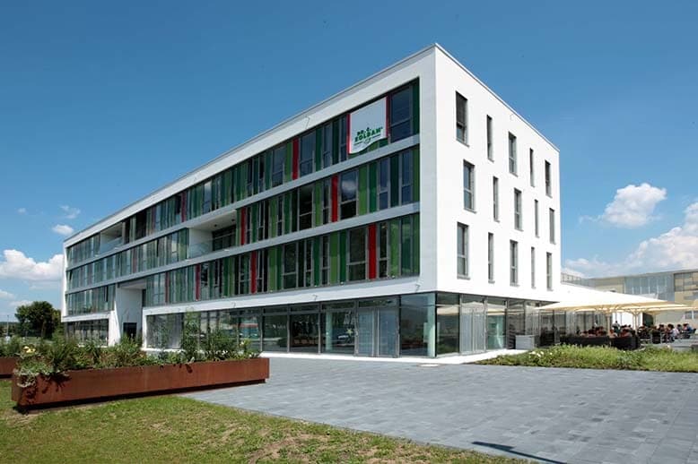 Building corner with green and red façade louvres, planters on the forecourt and a terrace with parasols