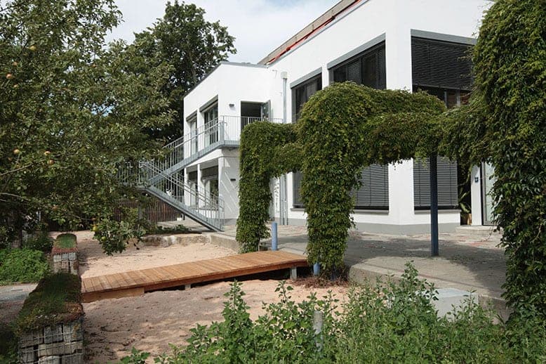 Außenbereich mit Sandfläche, Holzsteg und begrünter Pergola vor der weißen Fassade, links eine Außentreppe