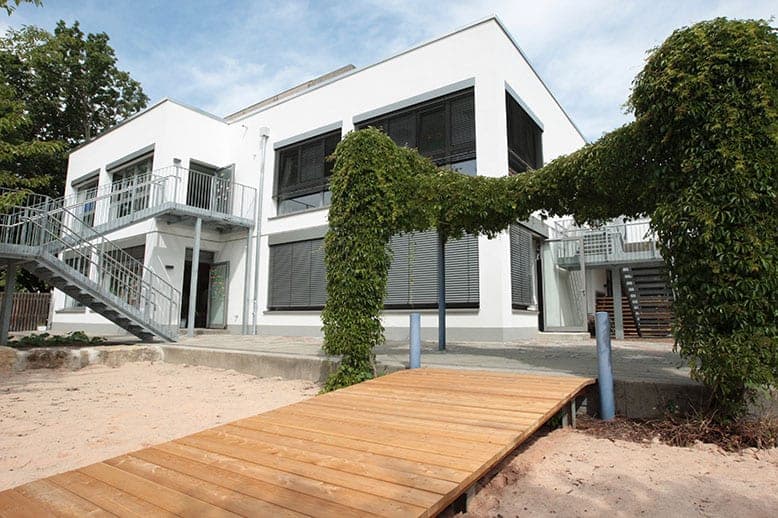 Blick über den Holzsteg zu den Außentreppen und der berankten Pergola am aufgestockten Kindergarten