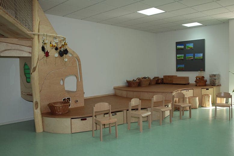 Gruppenraum mit Spielpodest aus Holz, Körben, Kinderstühlen und dem Kletterhaus am linken Rand