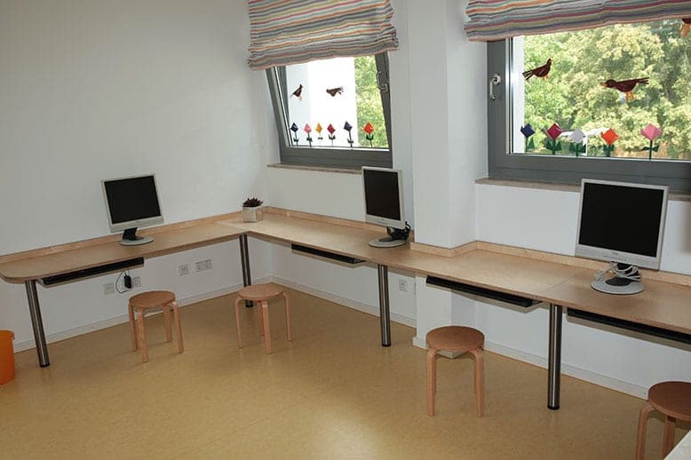 PC-Labor mit drei Bildschirmarbeitsplätzen an einer Holzplatte und Hockern unter den Fenstern