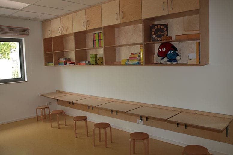 Wandregal mit Spielsachen über einer Reihe hochklappbarer Tische mit Hockern im Hort