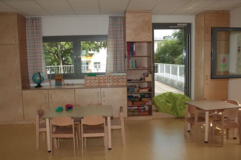 Gruppenraum mit Kindertischen, Holzschränken, Bücherregal und Glastür zum Balkon
