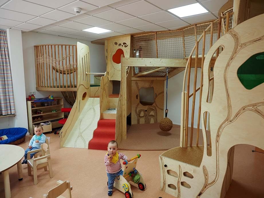 Spielraum mit zweigeschossiger Kletterlandschaft aus Holz, Rutsche, Netzgalerie und spielenden Kleinkindern