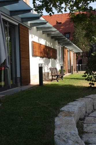 Garden side of the parish centre with glass canopy, timber louvres, a bench and a natural-stone edge in the lawn