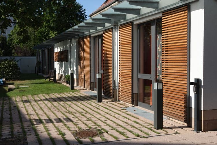 Sliding timber-louvre shutters along the façade with glass canopy, bollard lights and grass-joint paving