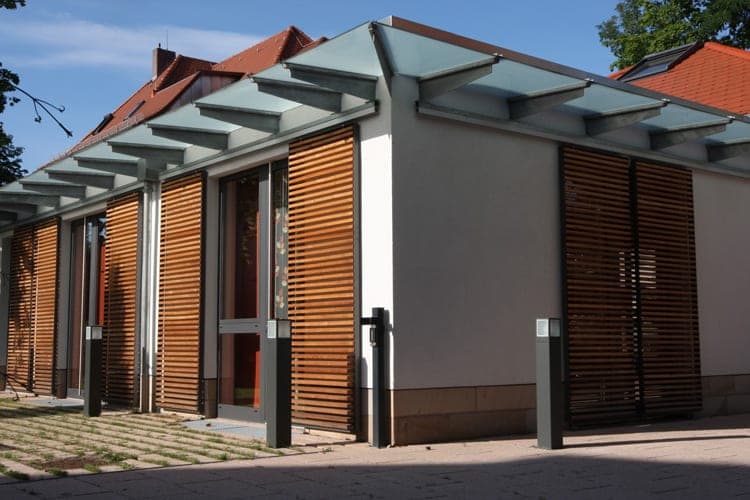 Corner view with a glass canopy on steel brackets, timber-louvre shutters and a stone plinth, tiled roofs behind