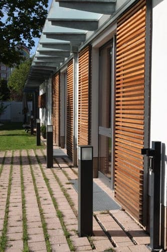 Perspective along the louvred façade beneath the glass canopy with a bollard light and grass-joint paving