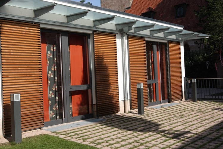 Glass doors with orange curtains and a leaf pattern between timber-louvre shutters beneath the glass canopy