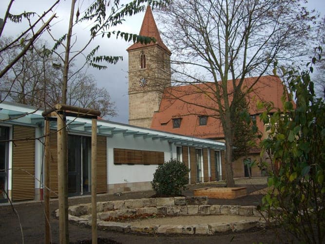 Parish centre with glass canopy in front of the sandstone tower of the Johanneskirche, a stone ring in the garden
