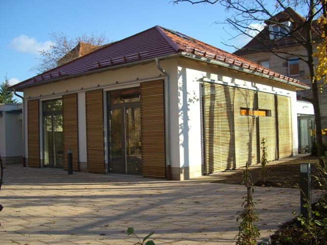 Oblique view of the pavilion with hipped roof and louvre wall with a window slit, young planting in front