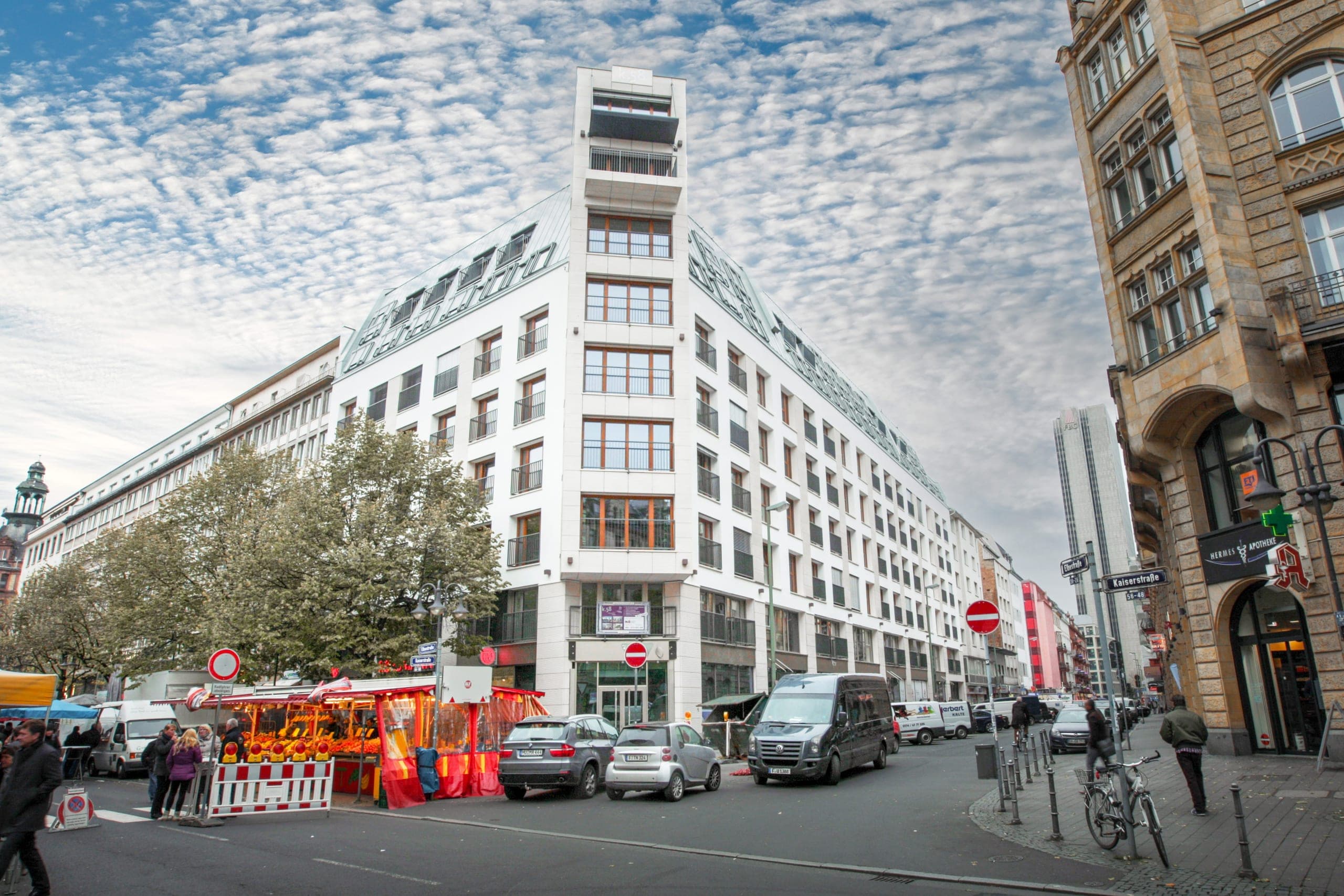 Realisiertes Eckgebäude im Bahnhofsviertel mit überhöhtem Eckturm, davor ein Marktstand und Passanten