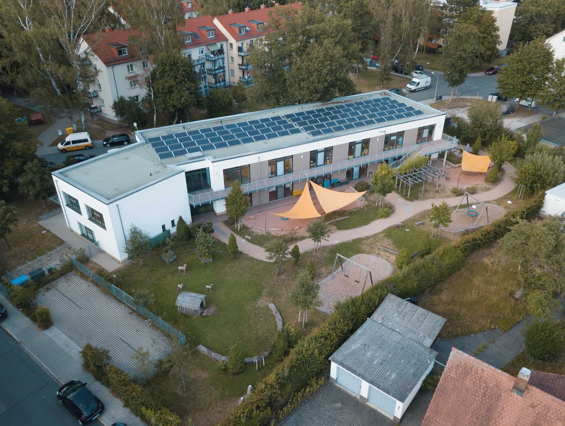 Aerial view of the two-storey day-care centre with photovoltaic roof, upper-floor gallery and play garden with sun sails