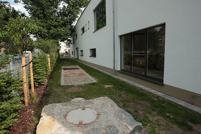 Side façade with large windows and a narrow lawn strip, with a stone block and round manhole cover in the foreground