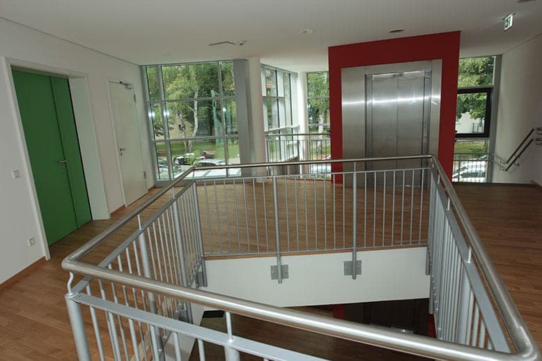 Upper-floor gallery of the foyer with metal railing, lift and glazed façade looking out onto greenery