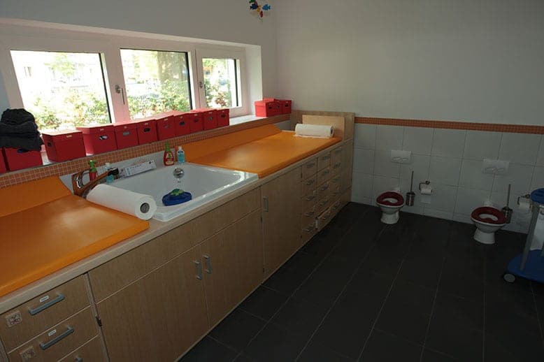 Nappy-changing area with orange mats, a basin and red storage boxes, with two children's toilets at the back