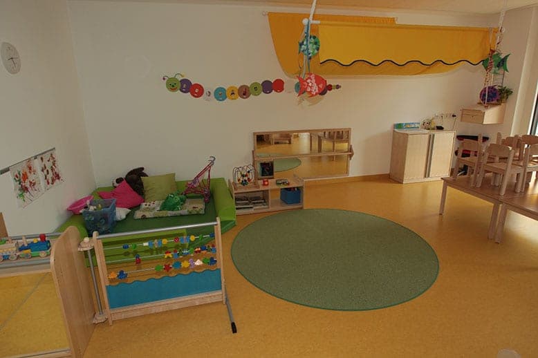 Group room with a green upholstered platform, mirror, round rug and children's tables on a yellow floor