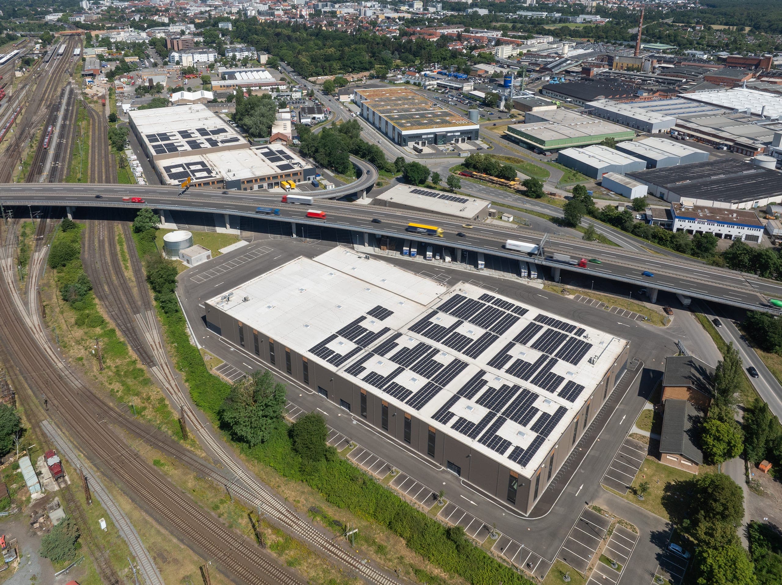 Schrägluftbild einer Halle mit Solardach zwischen Hochstraße und Gleisen, dahinter Gewerbebauten in Hanau
