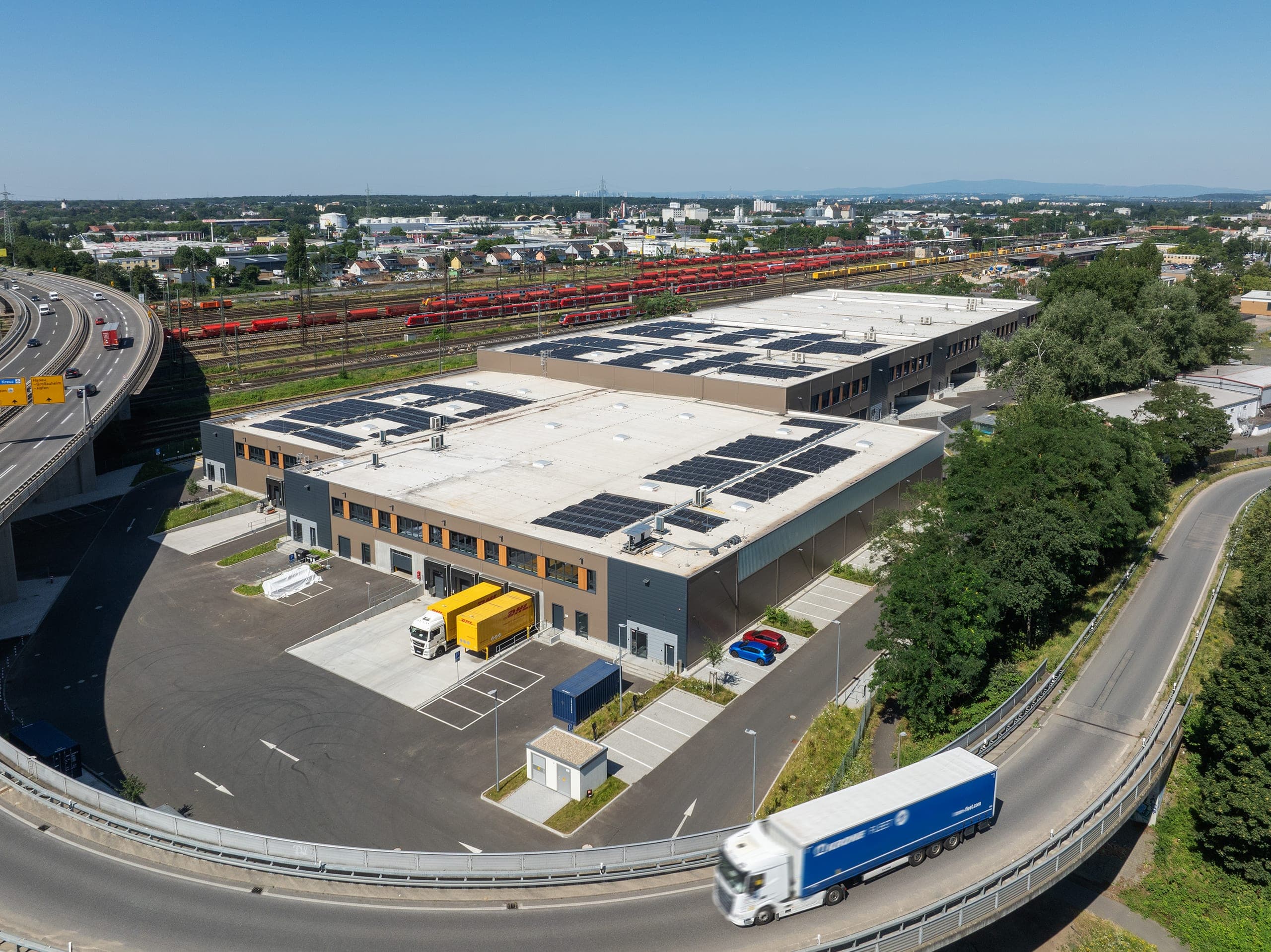 Luftbild einer Halle mit orangefarbenen Fensterrahmen und Lkw an der Anlieferung, dahinter Abstellgleise