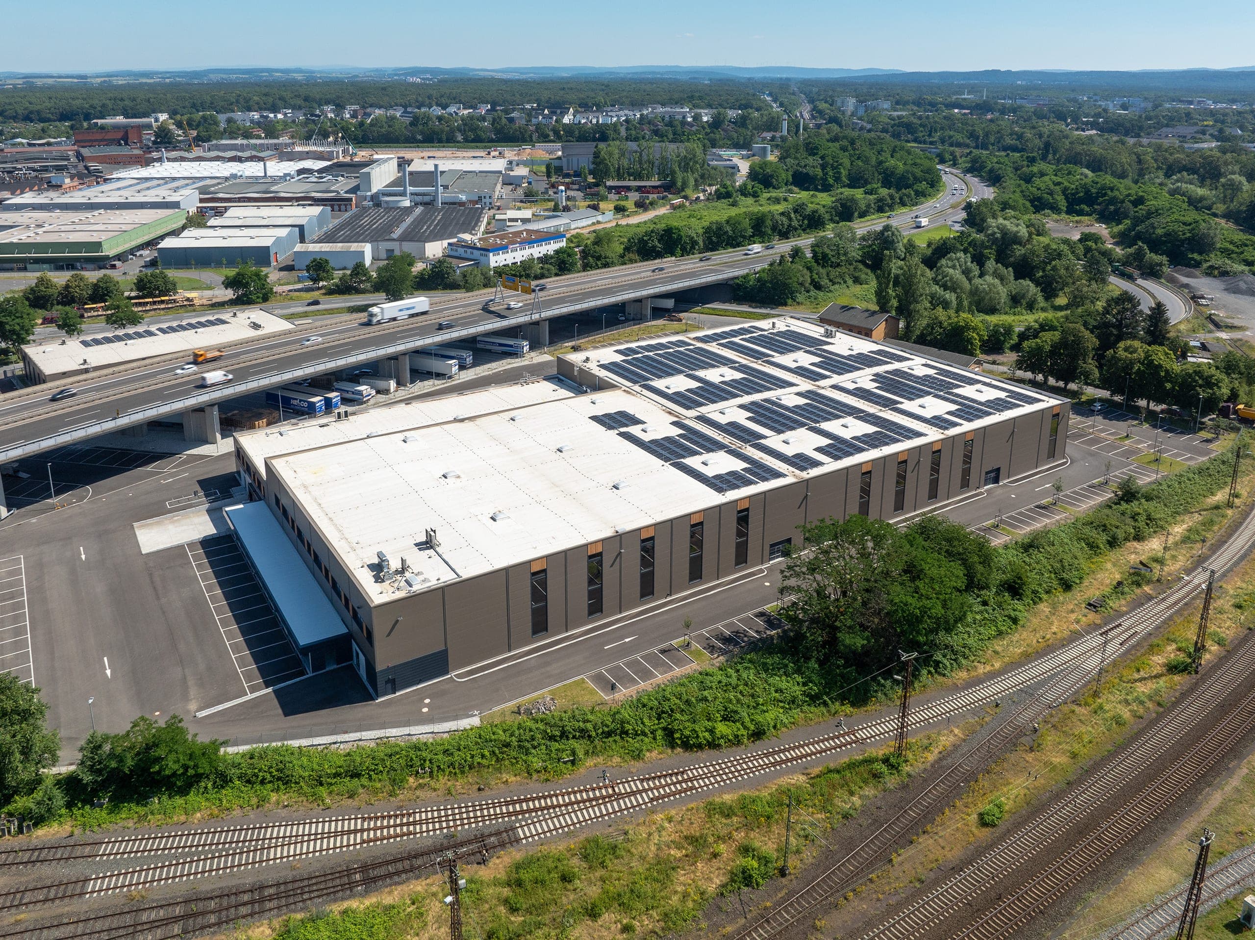 Luftbild der Halle mit Solarmodulen und Verladebereich, im Vordergrund Gleise, dahinter die Hochstraße