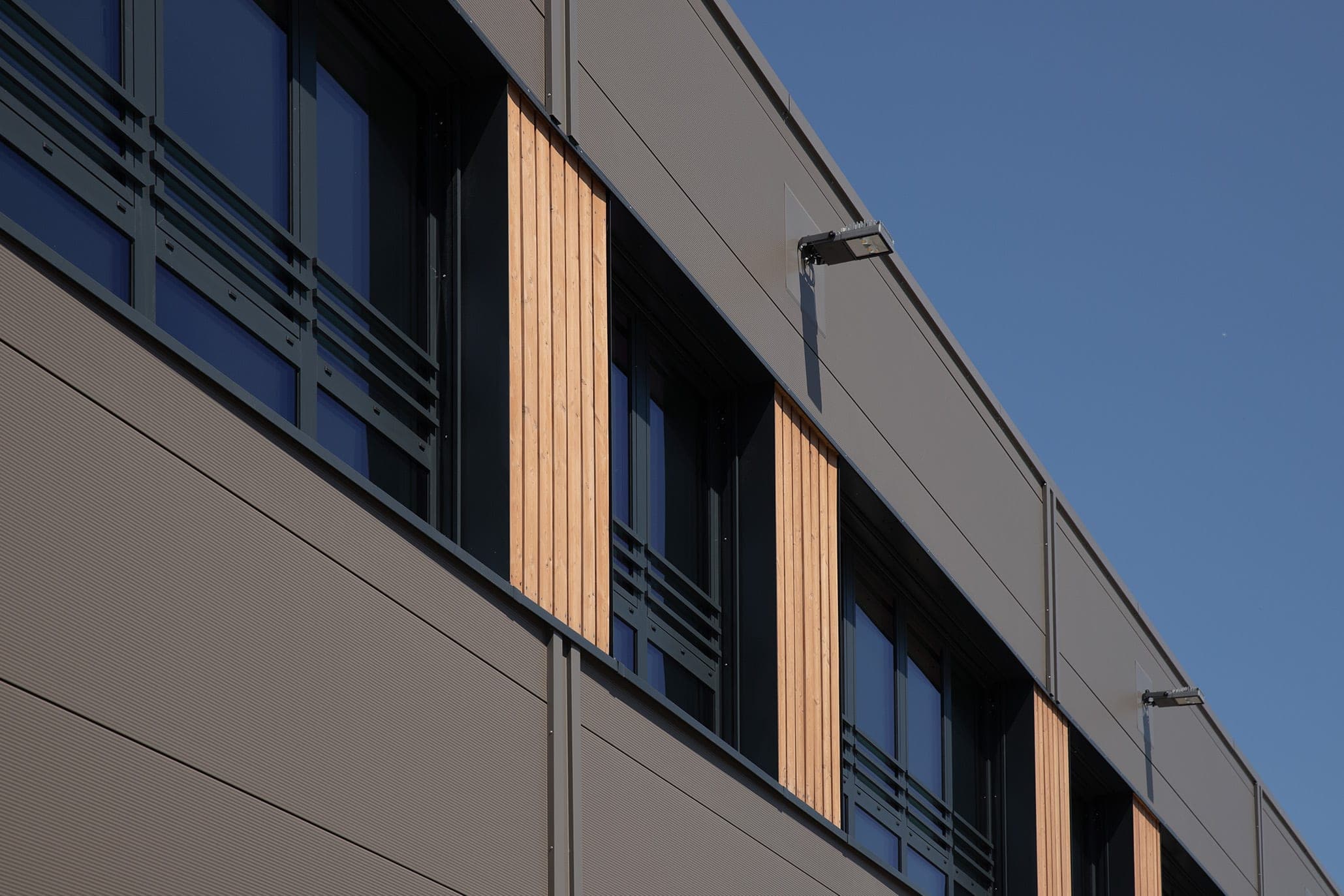 Nahaufnahme der Paneelfassade mit Holzlisenen zwischen Fensterbändern und Außenleuchten im Streiflicht