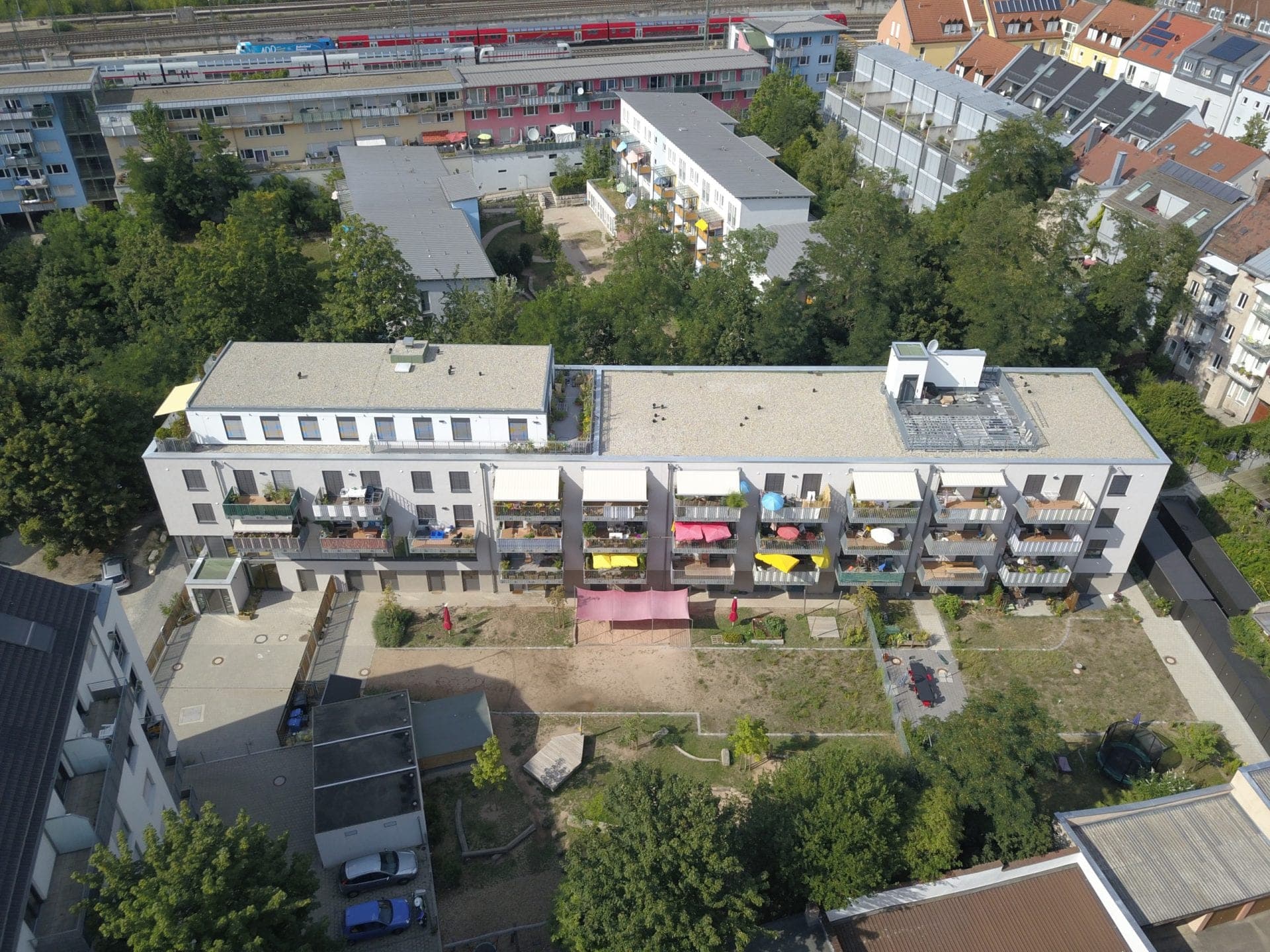 Luftbild der Balkonfassade mit Markisen und des Gartenhofs mit Sonnensegel, dahinter Bahngleise