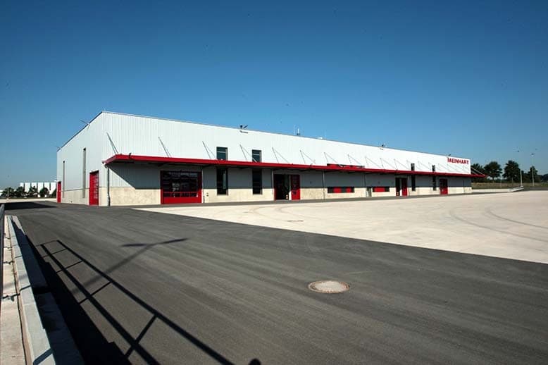 Logistics hall with a light metal façade, red doors and a red canopy band along the asphalt yard