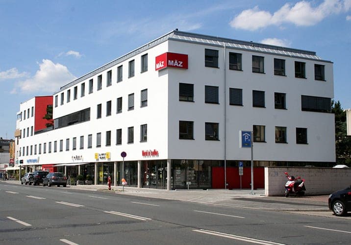Straßenansicht der weißen Gebäudeecke mit roter Wandfläche im Erdgeschoss, parkenden Autos und Motorroller